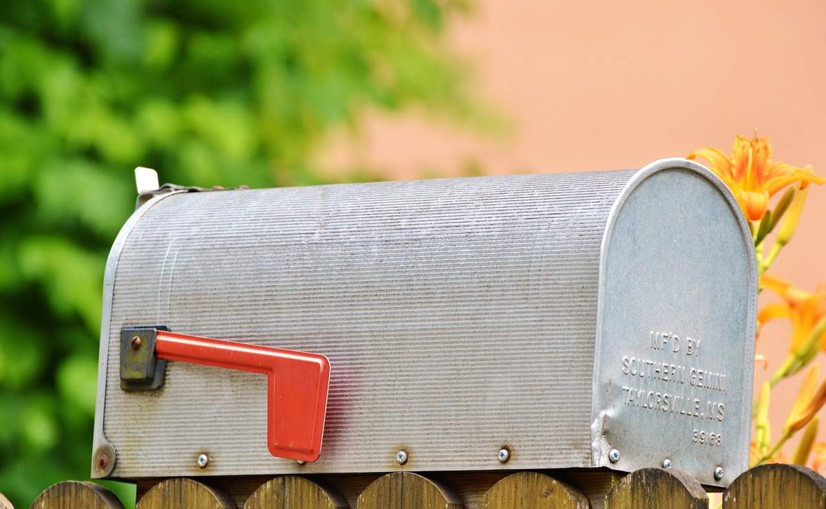 Boîte aux lettres métallique avec levier rouge sur clôture, fleurs jaunes